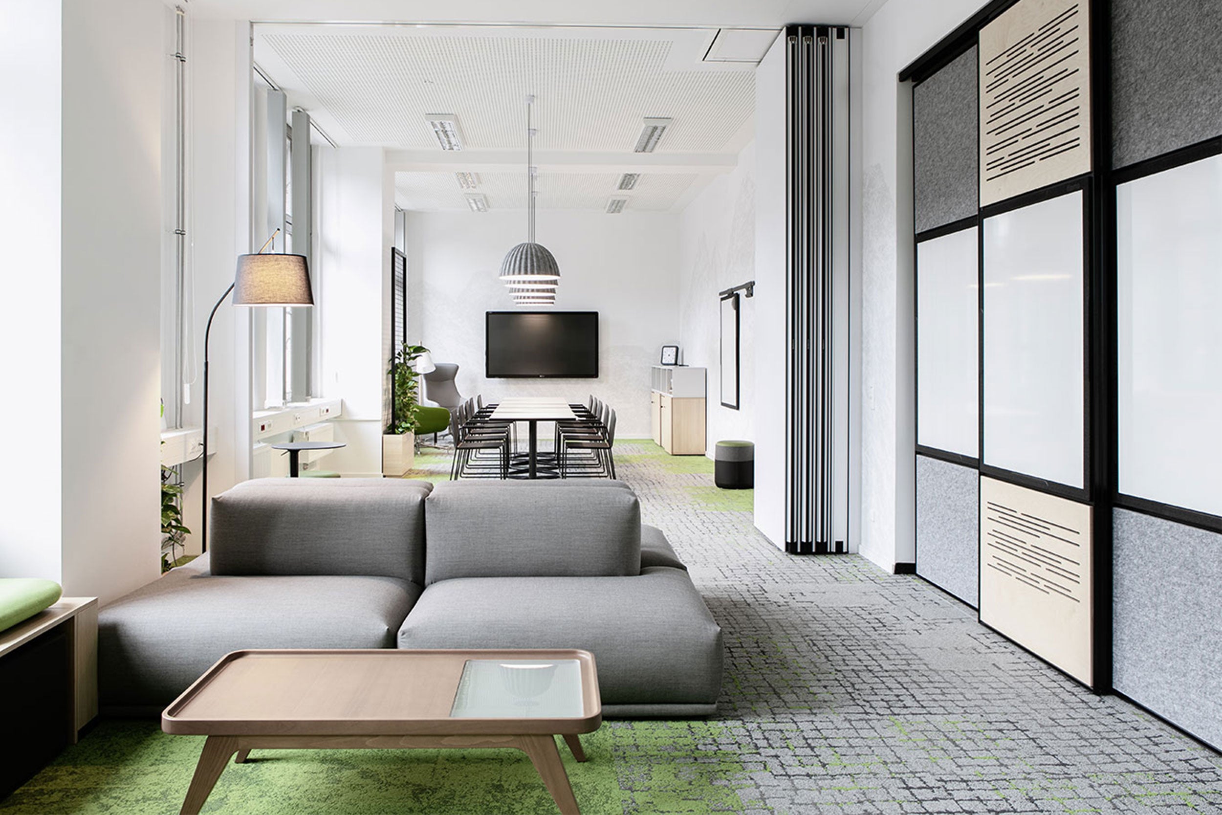Modern gestalteter Besprechungs- und Loungebereich mit grauem Sofa, Holz-Couchtisch, großem Konferenztisch, TV-Bildschirm und schallabsorbierenden Wand- und Deckenelementen in hellen Farben.